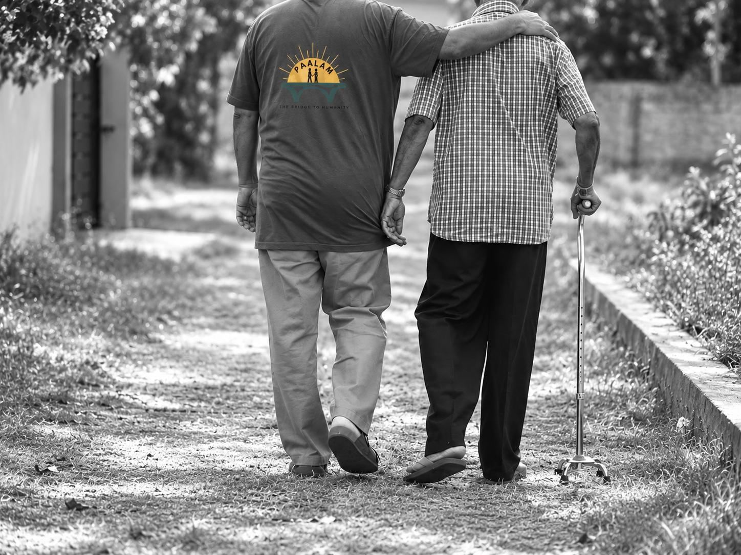 A volunteer wearing a Paalam T-shirt walking arm in arm with an older man using a walking stick